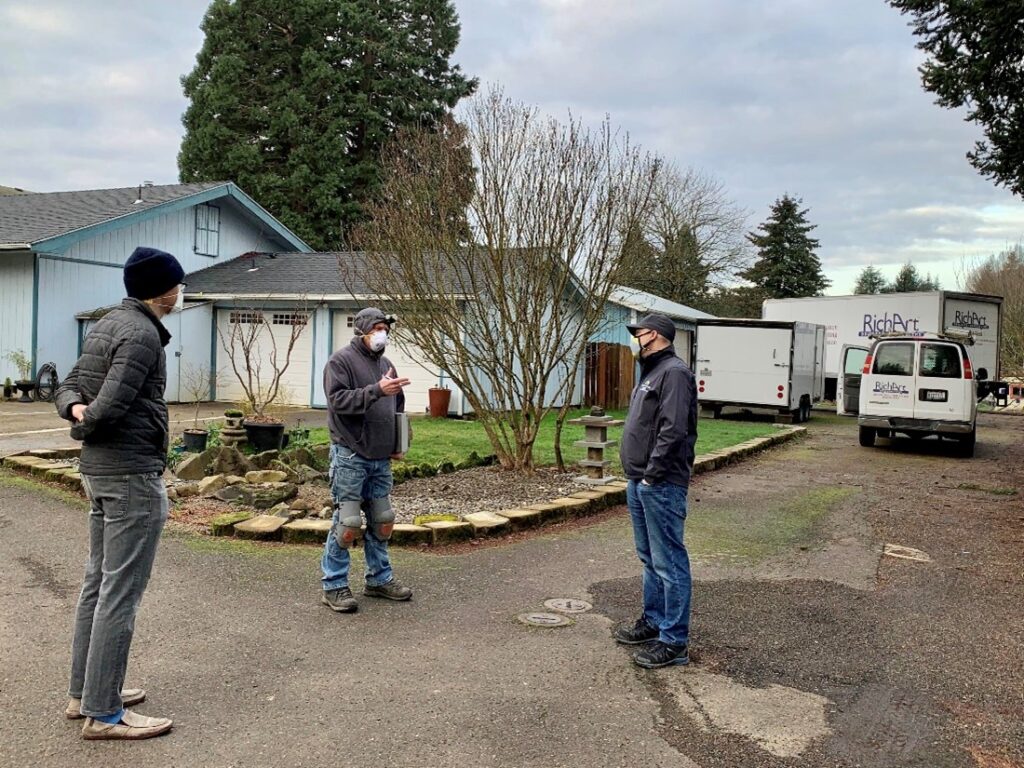 Three contractors stand six-feet apart facing each other in front of a residential home.
